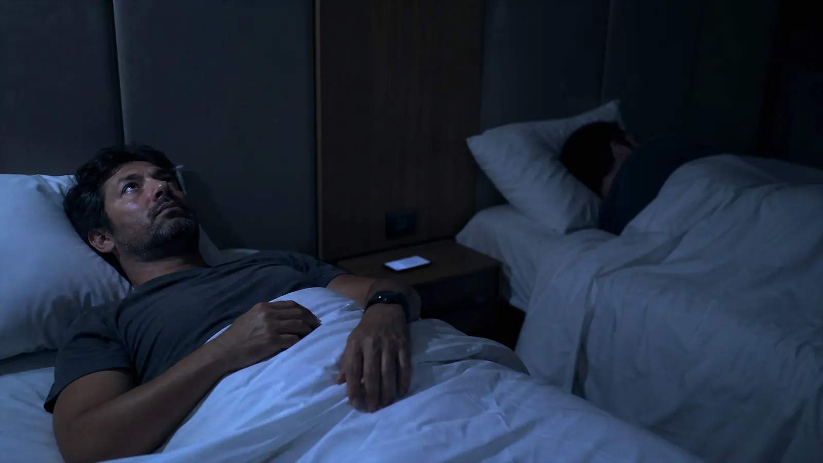 Traveler lying awake on a shared hotel bed while a roommate sleeps in the other bed